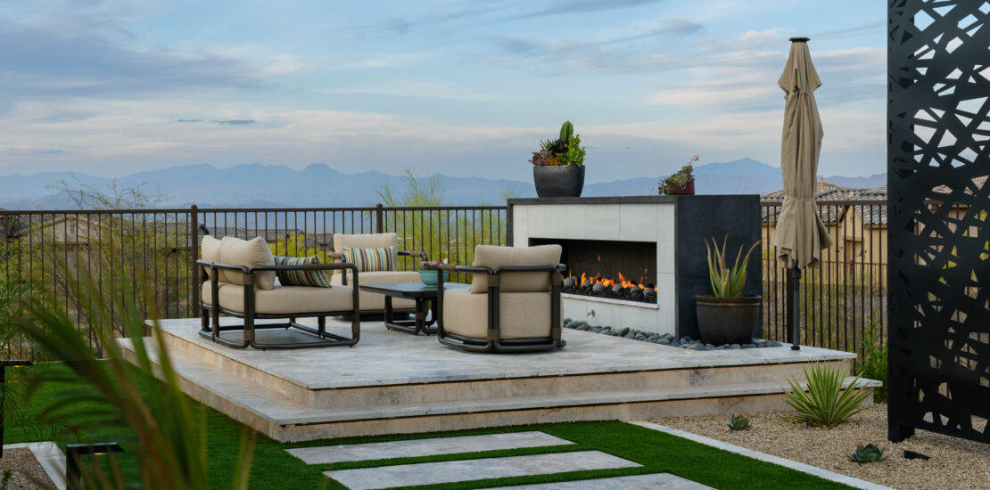 Image of backyard: hardscaping, turf, travertine steps, trellis in the background and raised firepit on a sun deck.