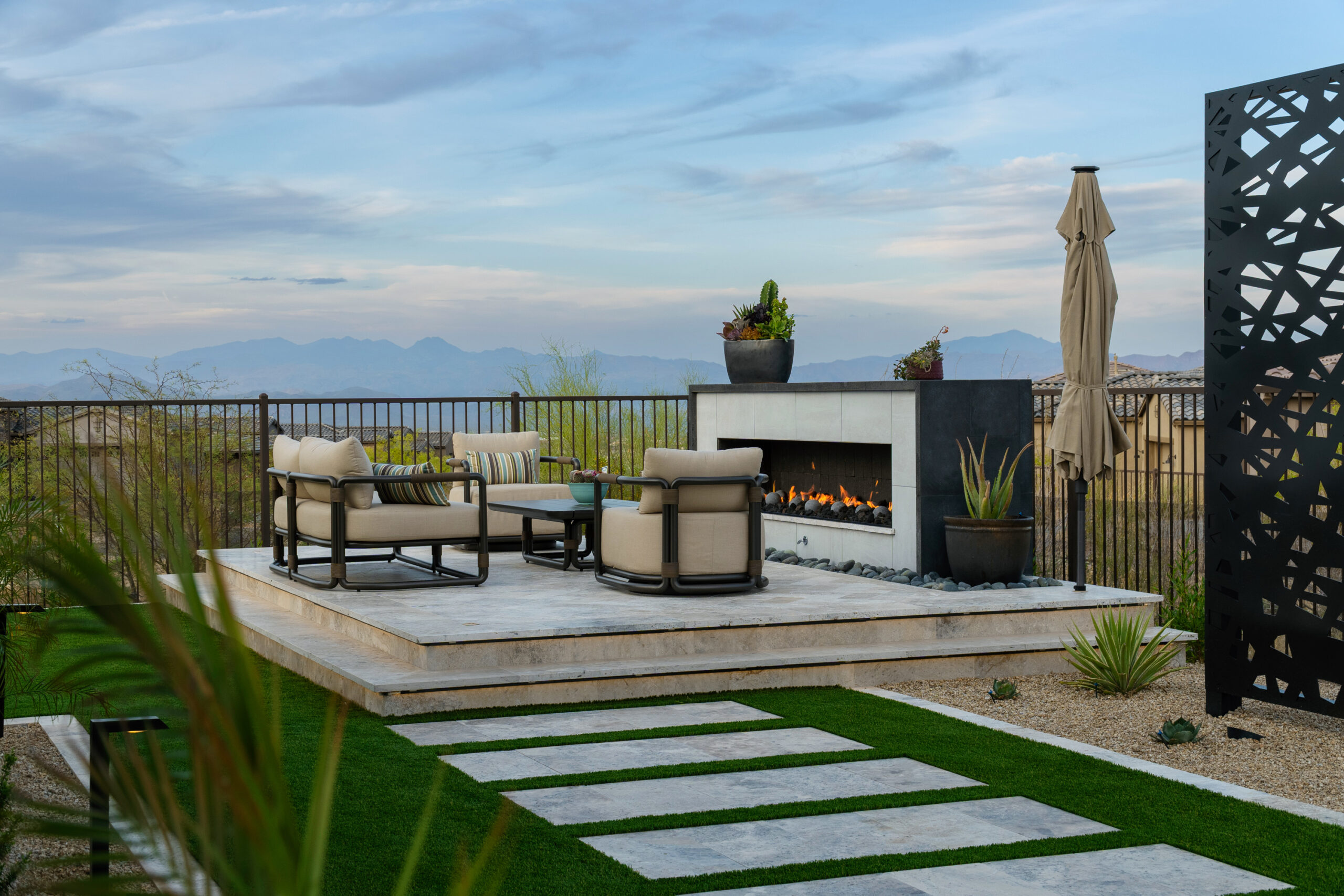 Image of backyard: hardscaping, turf, travertine steps, trellis in the background and raised firepit on a sun deck.