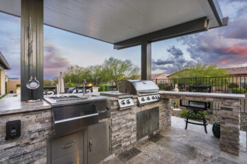 Custom outdoor kitchen with stacked stone finish, stainless grill, sink, and bar seating under a modern pergola by Kevens Landscape in Arizona.
