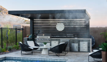 Alt text: Modern outdoor kitchen and BBQ area by Kevens Landscape featuring a stainless grill, pergola with misting system, and poolside seating in El Mirage, Arizona.